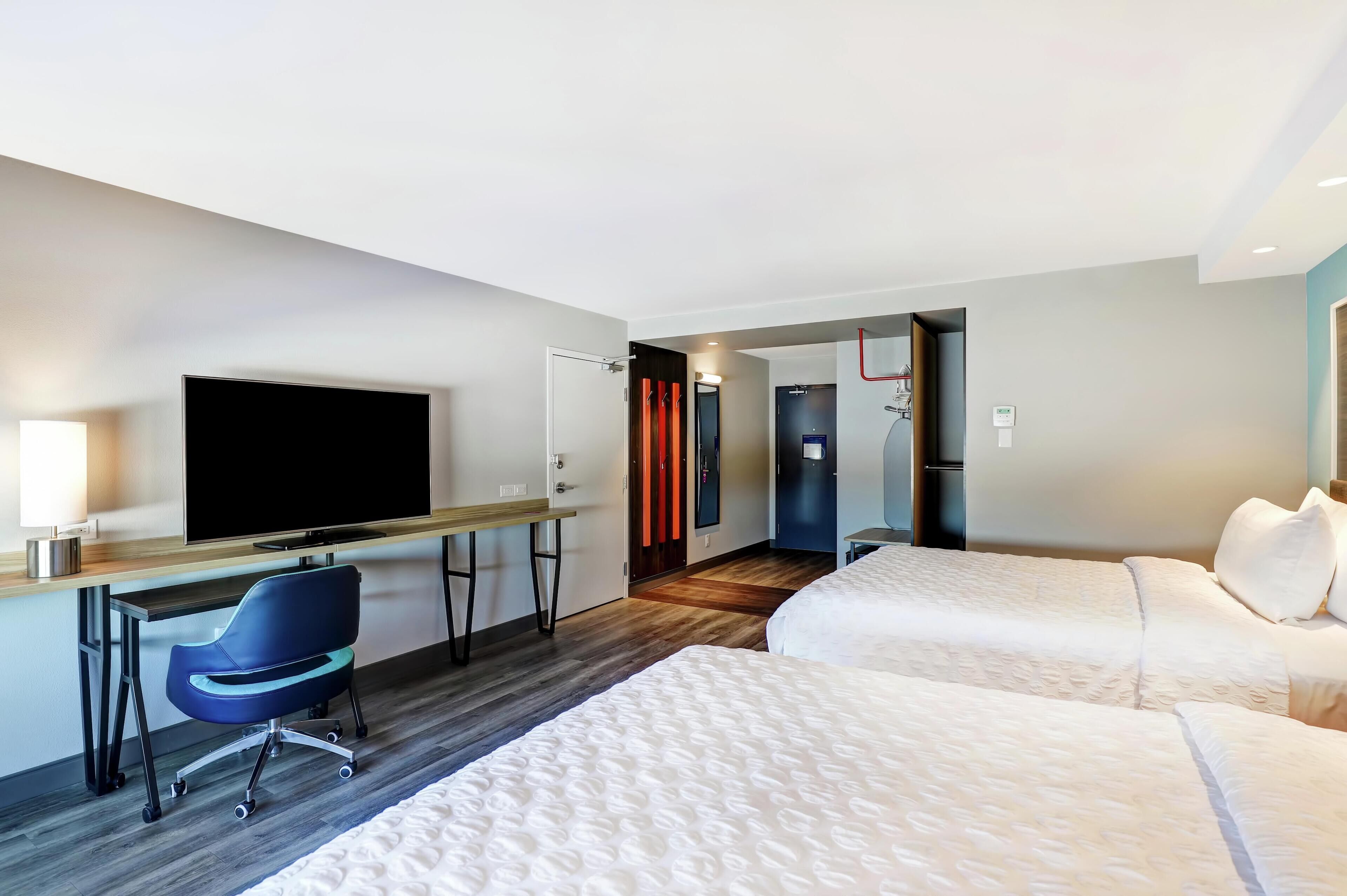 Habitación de hotel con dos camas, un escritorio con silla, un televisor de pantalla plana y una puerta que da a un baño.