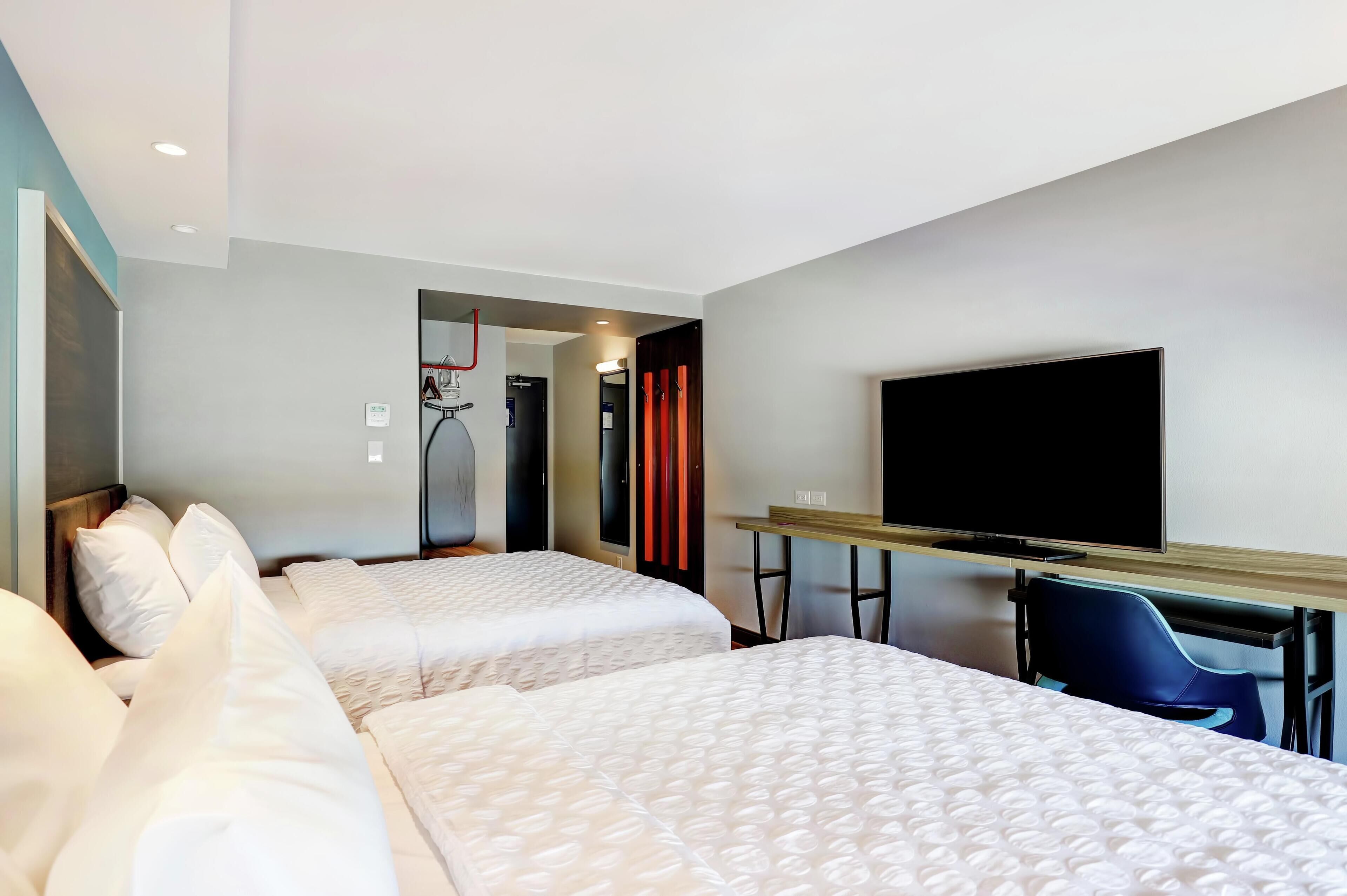 Habitación de hotel con dos camas, un escritorio con una silla, televisión y una cinta de correr al fondo.