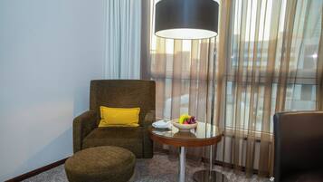 Ein Hotelzimmer mit braunem Sessel, gelbem Kissen, rundem Tisch und einer Obstschale sowie einer Lampe.