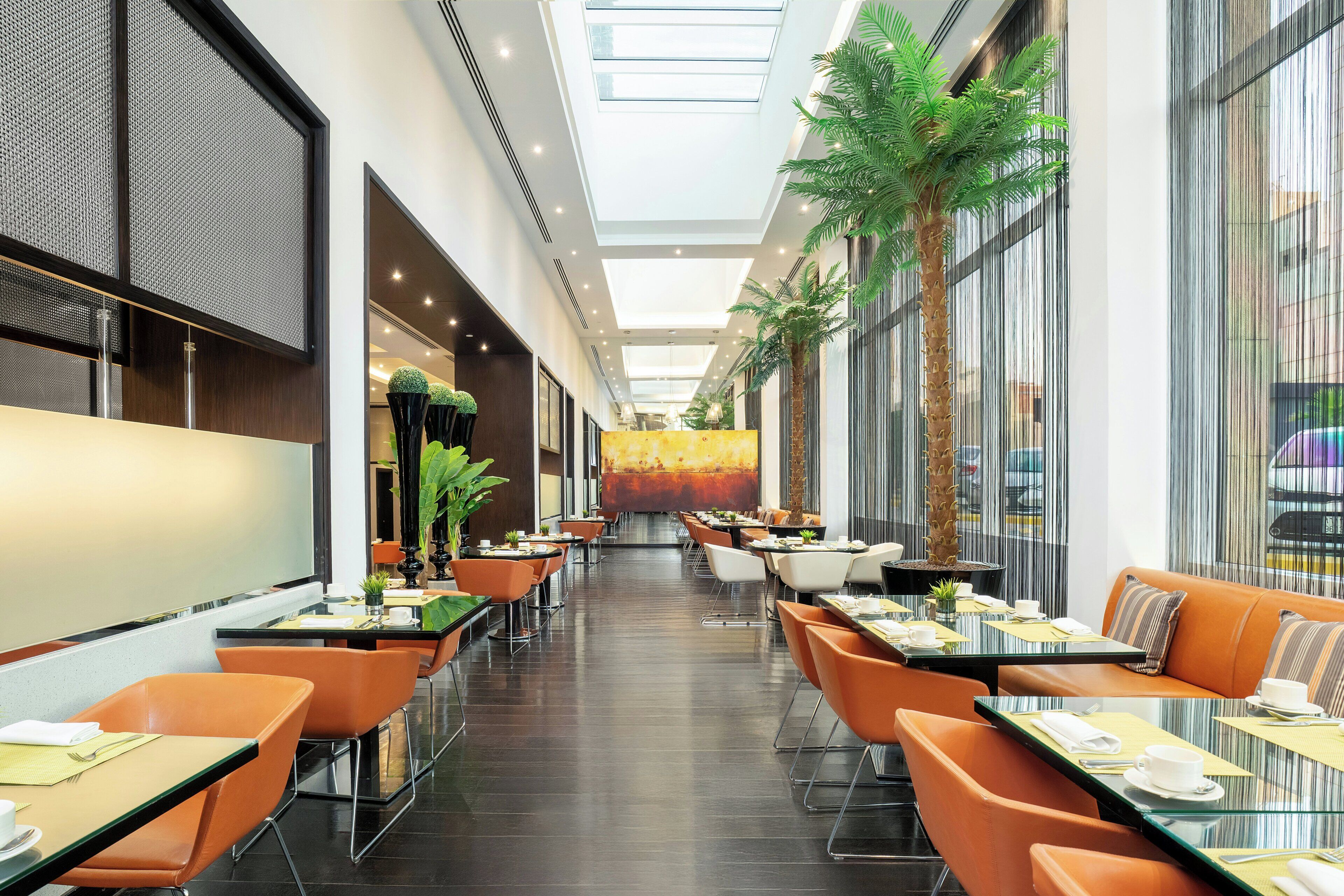 A modern hotel corridor with tables and chairs, a central art piece, and large windows.