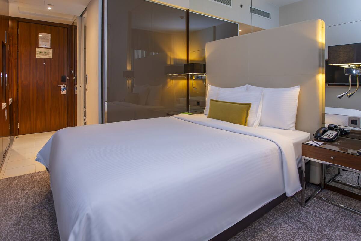 Hotellihuone, jossa on suuri sänky, yöpöytä, jossa on puhelin ja kello, peili ja ovi kylpyhuoneeseen.