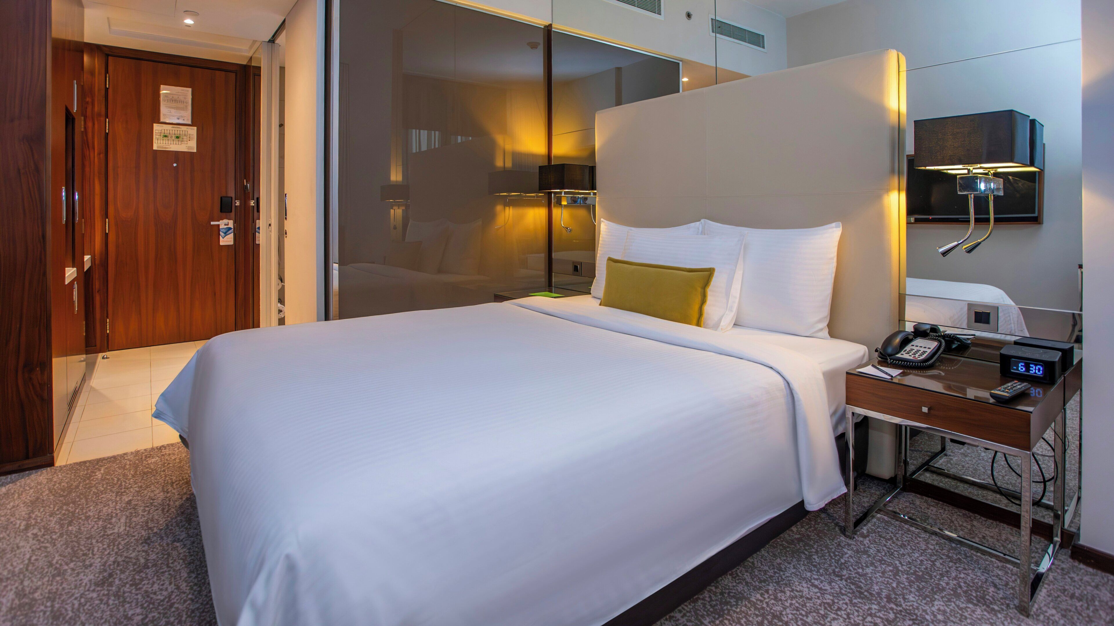 Ein Hotelzimmer mit einem großen Bett, einem Nachttisch mit Telefon und Wecker, einem Spiegel und einer Tür mit Holzrahmen.