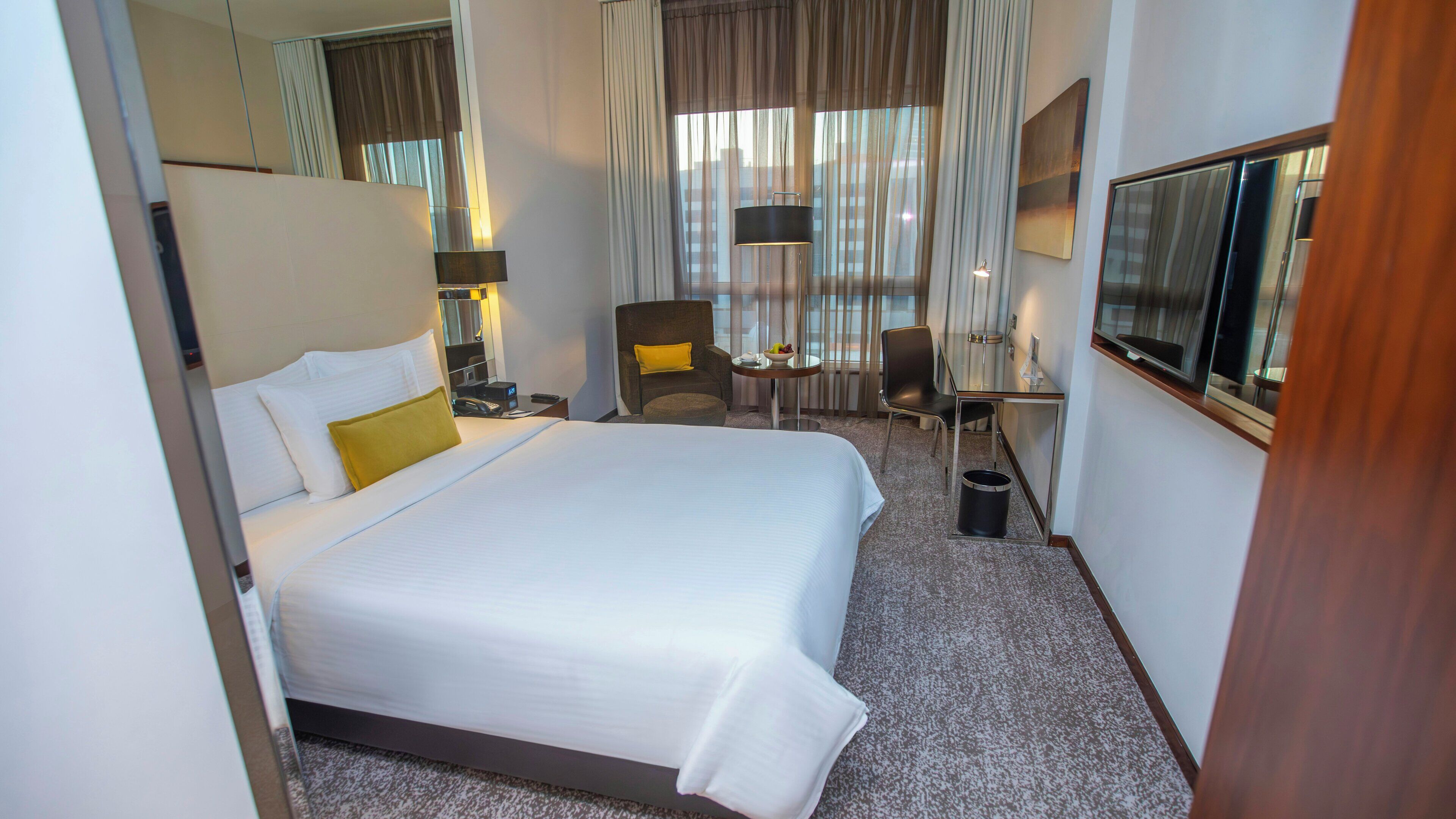 A hotel room with a large bed, a desk, a chair, a TV, and a window with curtains.