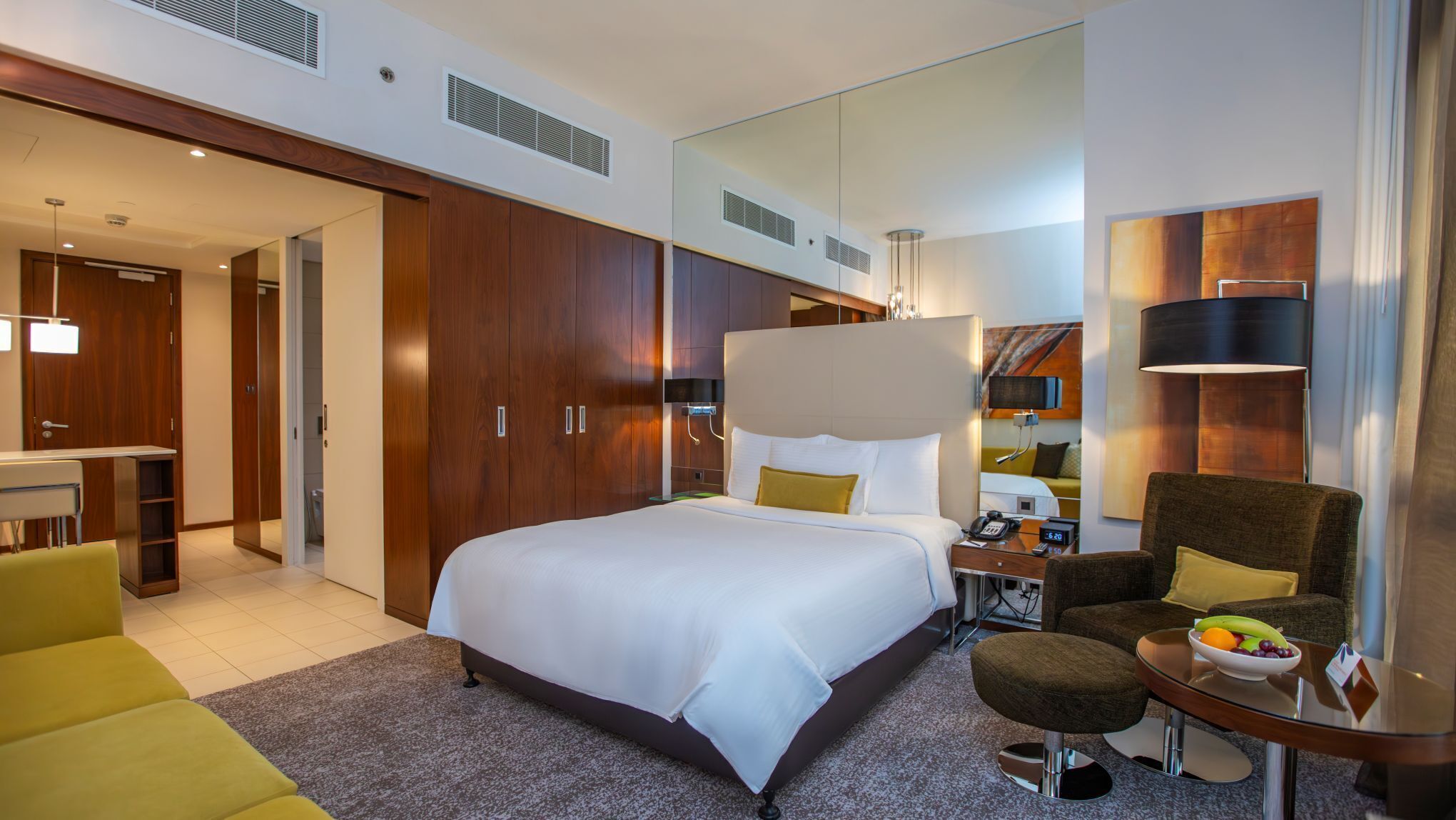 A hotel room with a large bed, a desk with a telephone, a chair, a small table with a fruit bowl, and a mirror.