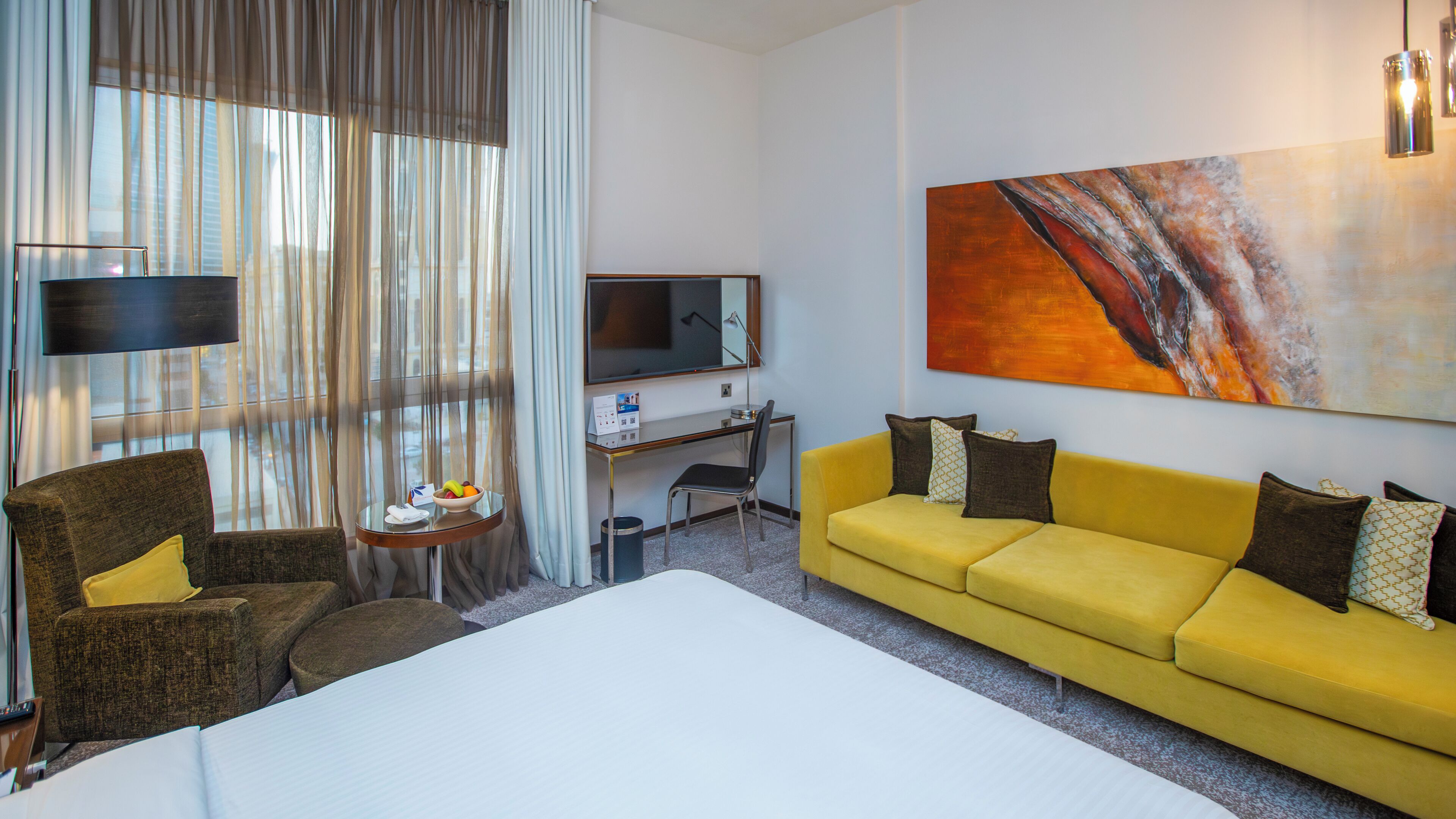 Habitación de hotel con una cama grande, un sofá amarillo, un escritorio con computadora, televisor y un cuadro en la pared.