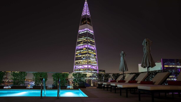 Vista nocturna de un edificio alto e iluminado con una aguja, piscina y sillones de relax con sombrillas.