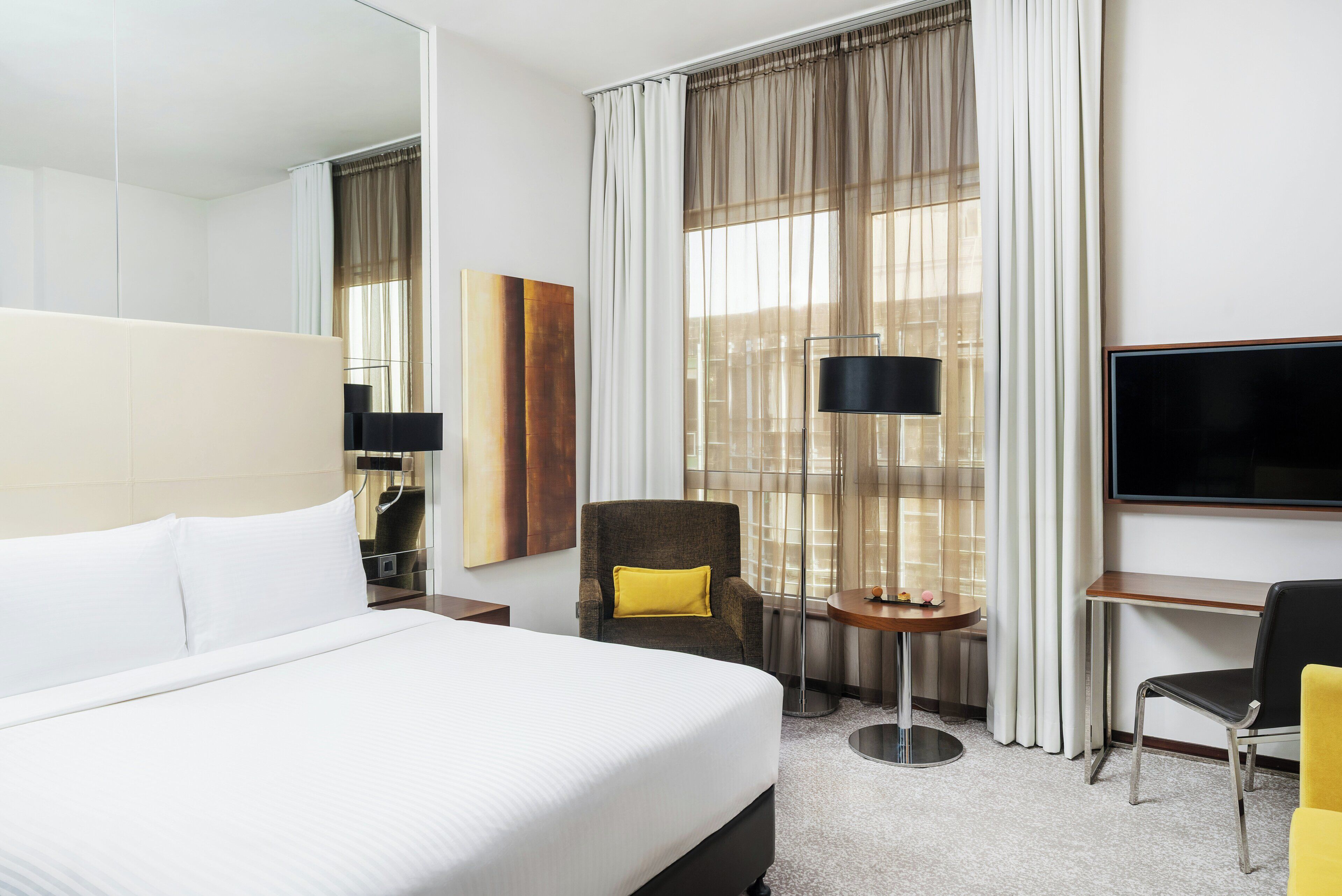 Une chambre d’hôtel avec un lit, une chaise, un bureau, une télévision et une fenêtre avec des rideaux.