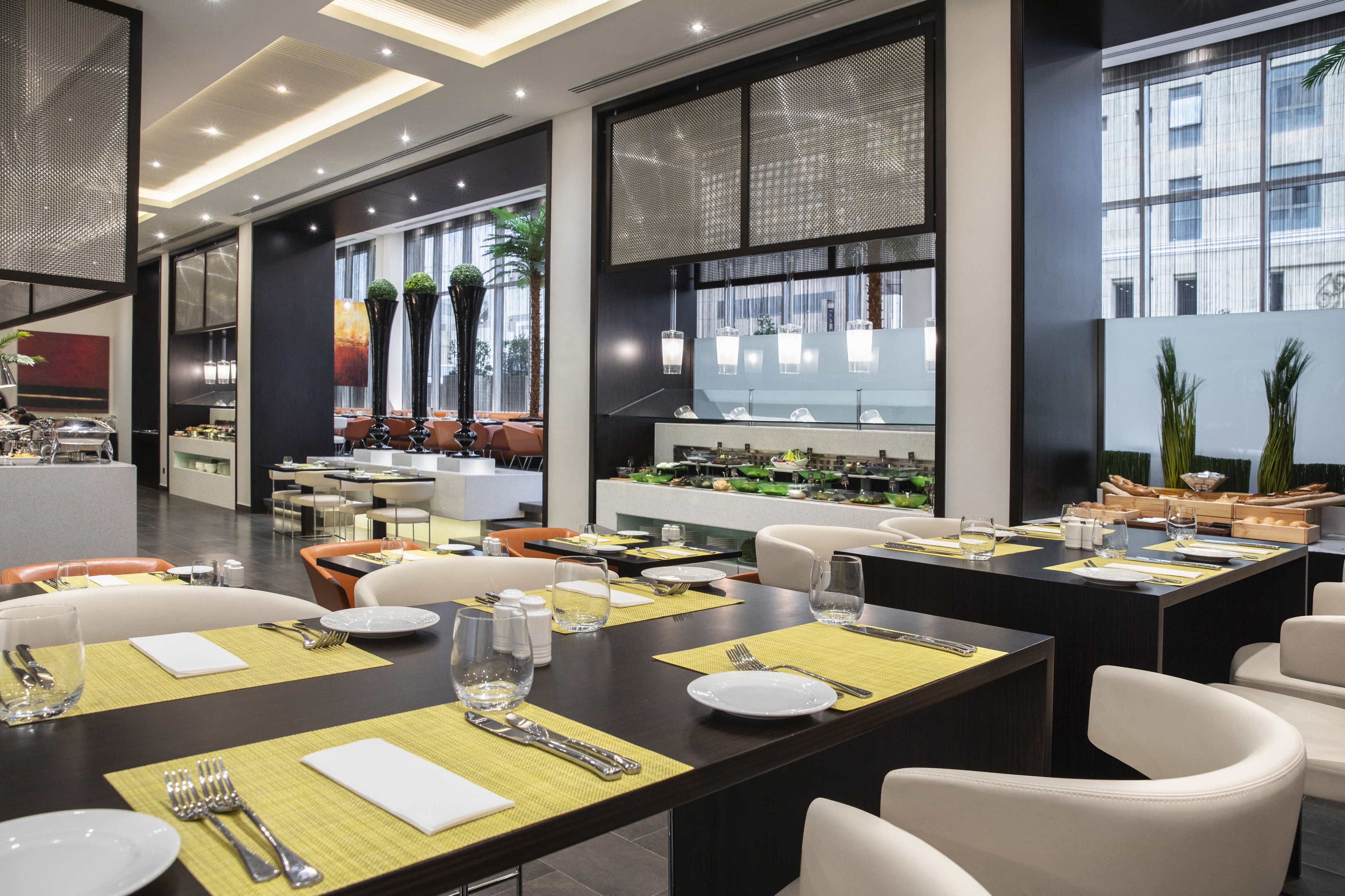 A modern dining area with multiple tables set for meals, large windows offering views of the city, and a variety of seating options including chairs and sofas.