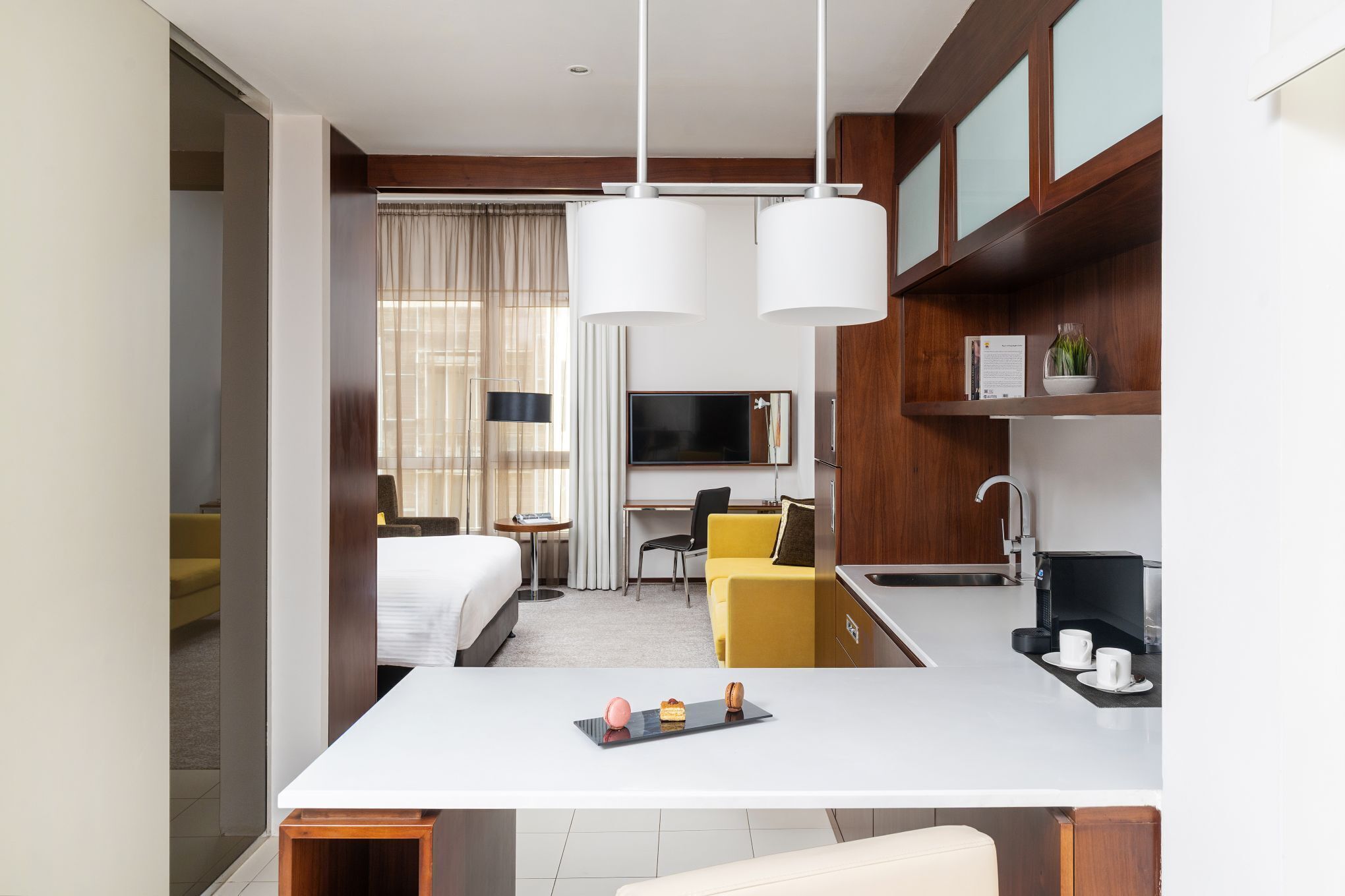A modern hotel room with a bed, a desk, a chair, a small table with a tray of pastries, and a kitchenette area.