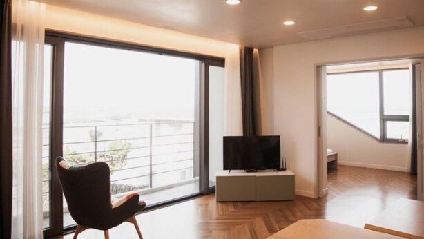 A modern living room with large windows, a flat-screen TV on a low cabinet, and a wooden floor. - Sinchon Blues (Jeju City)