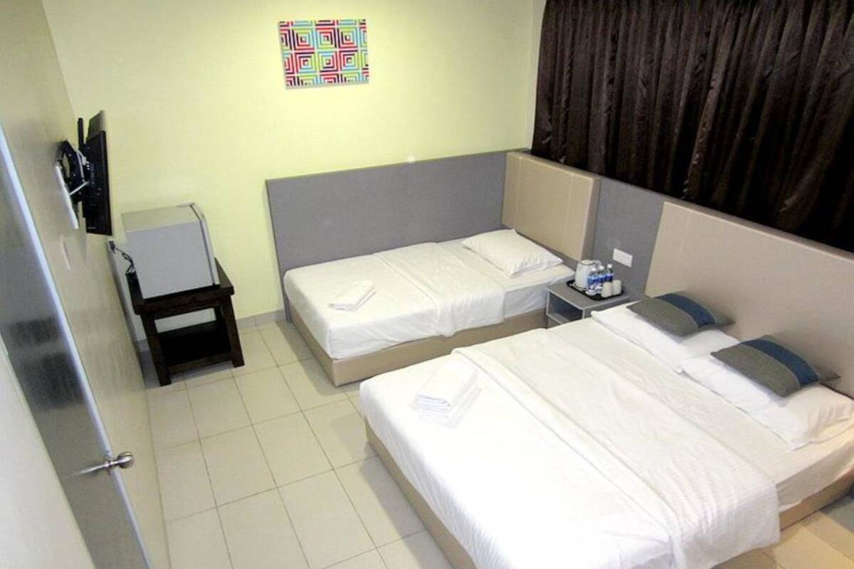 A hotel room with two single beds, a small desk with a chair, a TV mounted on the wall, and a colorful artwork above the bed.