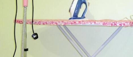 A pink vacuum cleaner, an ironing board with a floral-patterned edge, and a blue iron on a tiled floor.