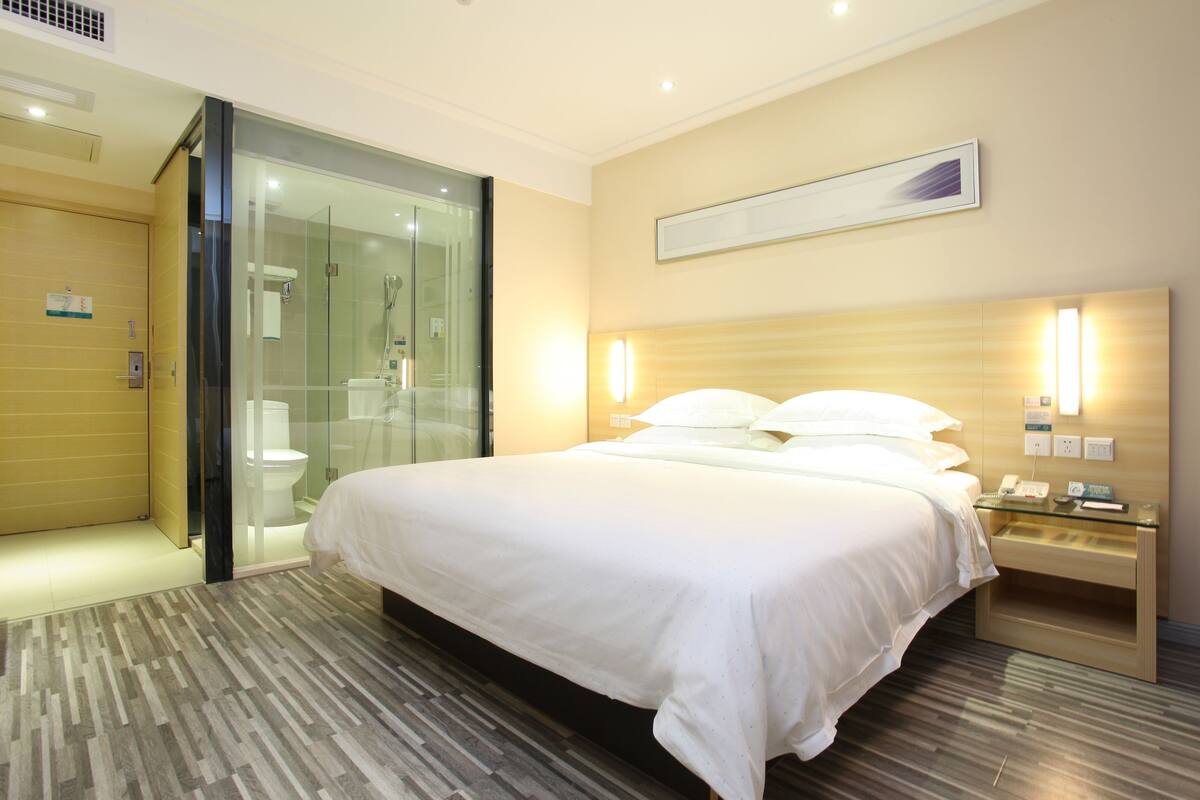 A modern hotel room with a large bed, a glass shower enclosure, and a bedside table with a phone and a lamp.