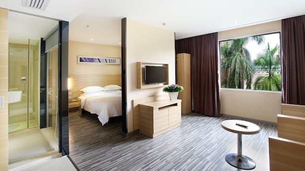 Ein modernes Hotelzimmer mit Bett, Fernseher, kleinem Tisch und großem Fenster mit Blick auf Palmen.