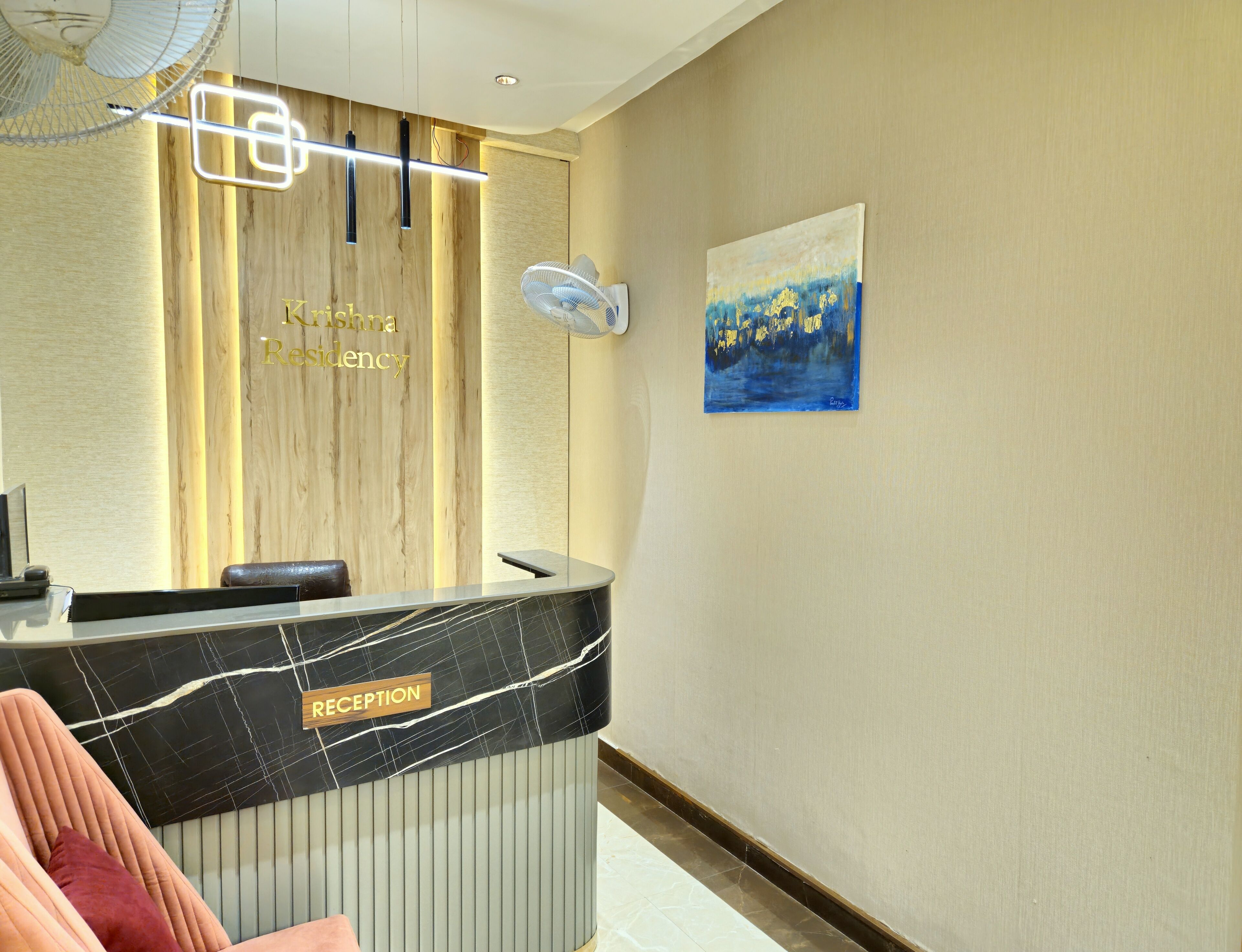A reception desk with a marble top, a wooden wall with a sign reading "Krishna Residency," a painting on the wall, and a fan on the ceiling.