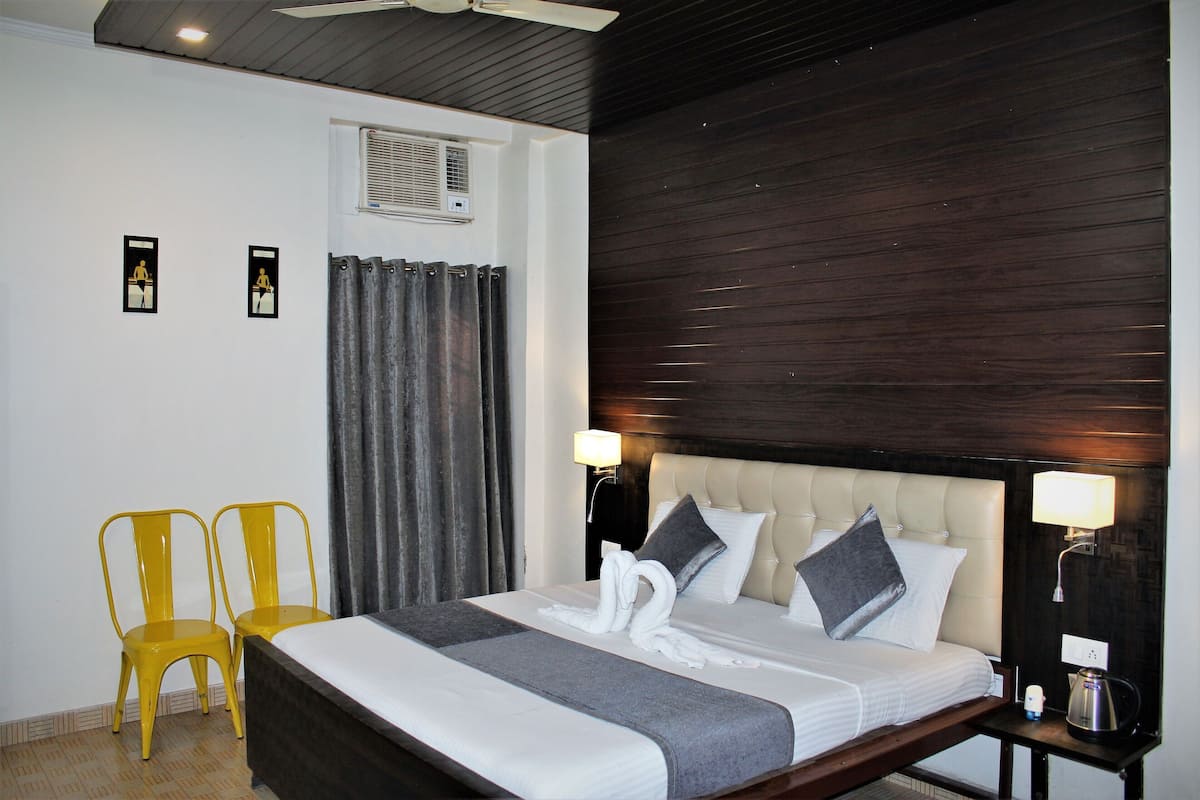 A hotel room with a bed, two chairs, a nightstand, a lamp, and an air conditioning unit.
