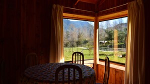 Una cabaña de madera con una mesa de comedor y sillas, ventanas grandes con cortinas, y vista a un paisaje montañoso.
