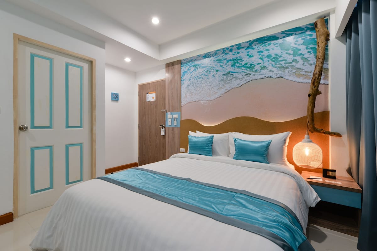 A hotel room with a large bed, a nightstand with a lamp, a wall mural of a beach scene, and a door with a sign.