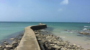 Un chemin de béton mène à une côte rocheuse bordée d’une eau d’un bleu limpide et à une île située au loin.