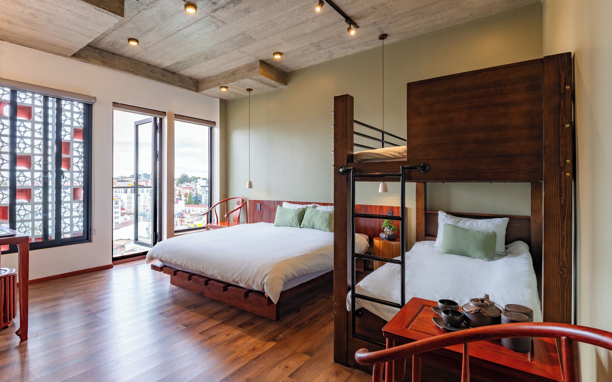 Ein Zimmer mit zwei Betten, einem Holztisch und Blick auf die Stadt durch große Fenster.