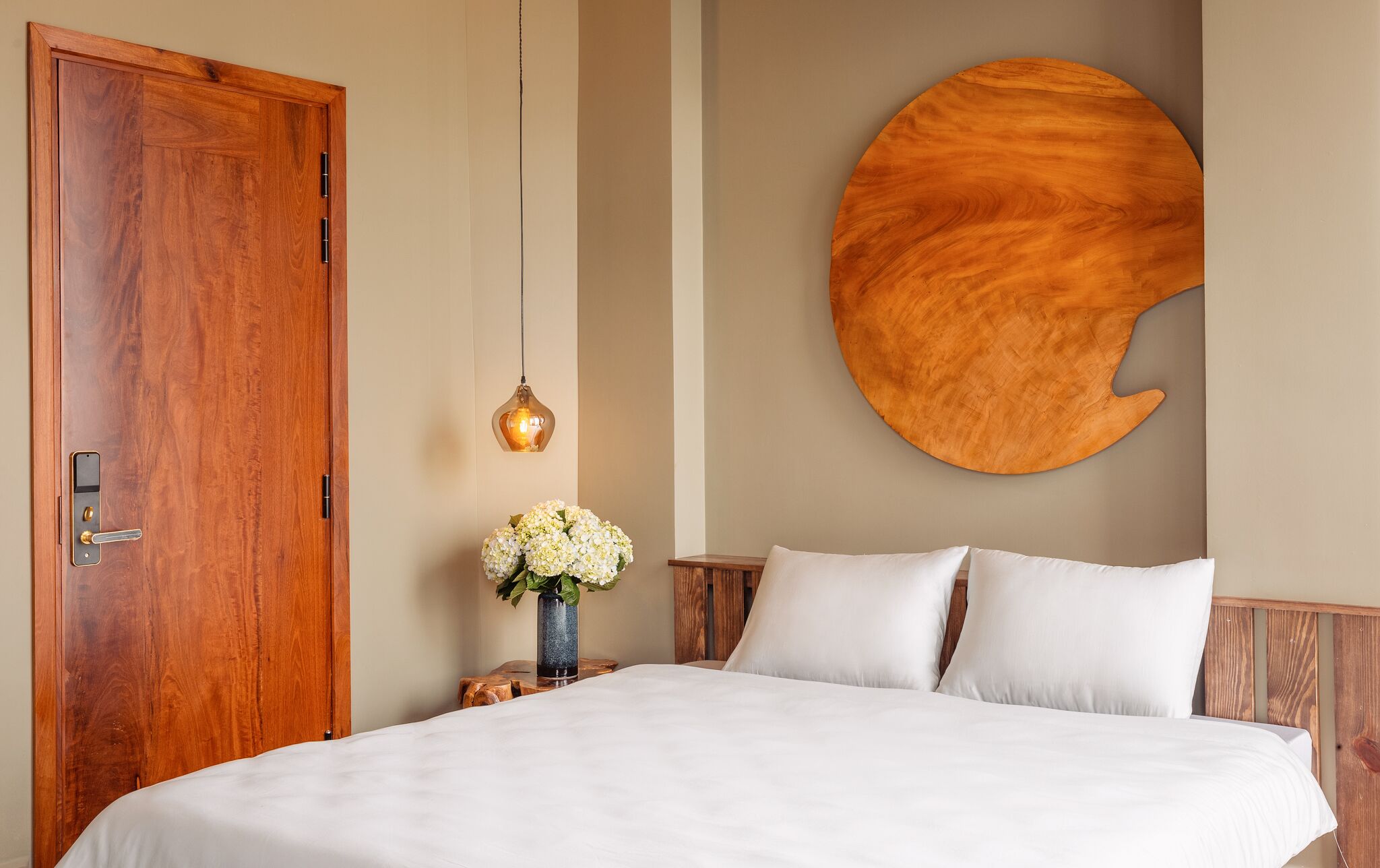 A bedroom with a wooden door, a round wall art piece, a bedside table with a vase of flowers, and a bed with white bedding.