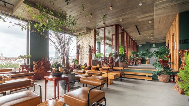 Amplio espacio interior con bancos y mesas de madera, grandes ventanales con vistas a un paisaje urbano y diversas plantas en macetas.