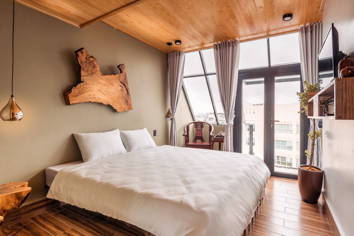 Une chambre à coucher moderne avec un grand lit, un plafond en bois, une grande fenêtre avec des rideaux, une tête de lit en bois, une table de chevet et une plante en pot.