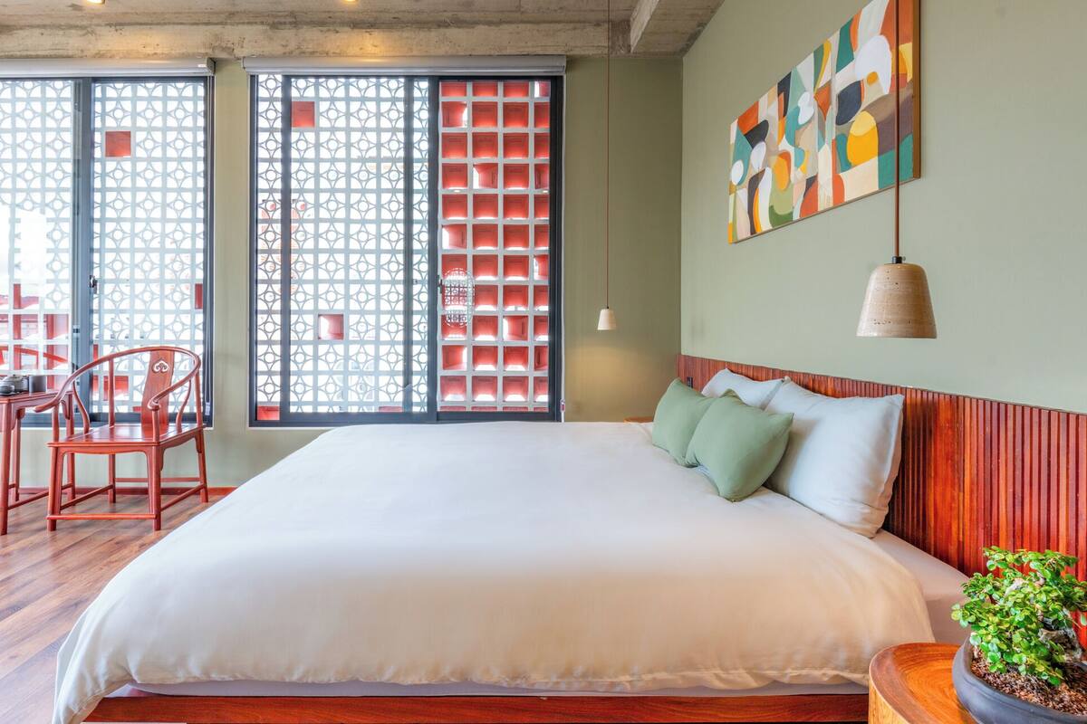 Une chambre d’hôtel avec un grand lit, une tête de lit en bois, une plante en pot et une fenêtre agrémentée d’un paravent décoratif.
