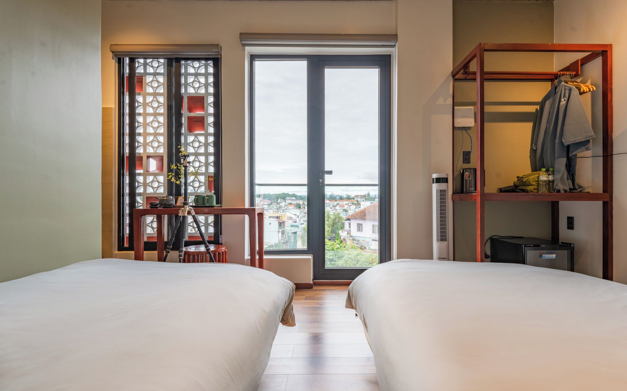 Een hotelkamer met twee bedden, een balkon met uitzicht op de stad, een houten plank met spullen en een bureau met een lamp.
