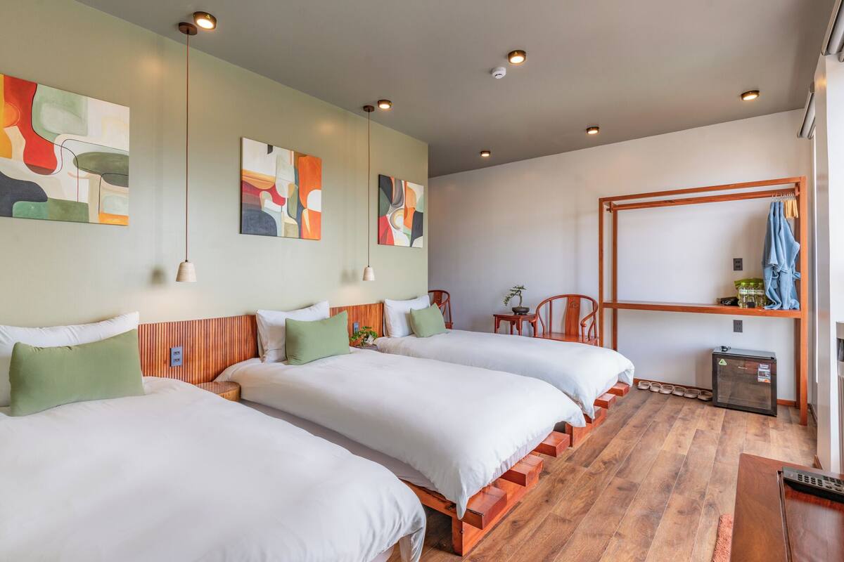 Une chambre d’hôtel avec deux lits, des têtes de lit en bois, des œuvres d’art abstraites accrochées au mur, une étagère en bois avec des serviettes et une petite table sur laquelle est posée une plante.