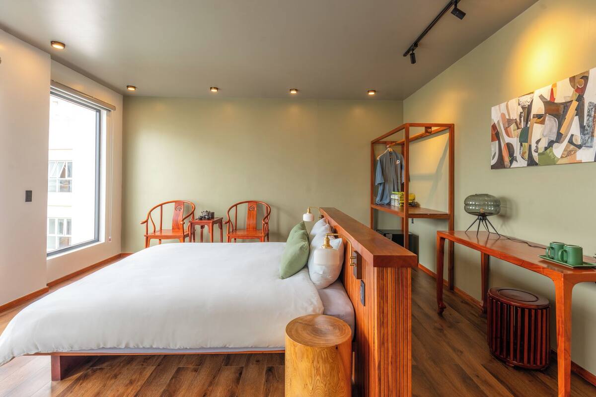 Une chambre à coucher avec un grand lit, deux chaises en bois, une petite table, une étagère suspendue et un tableau au mur.