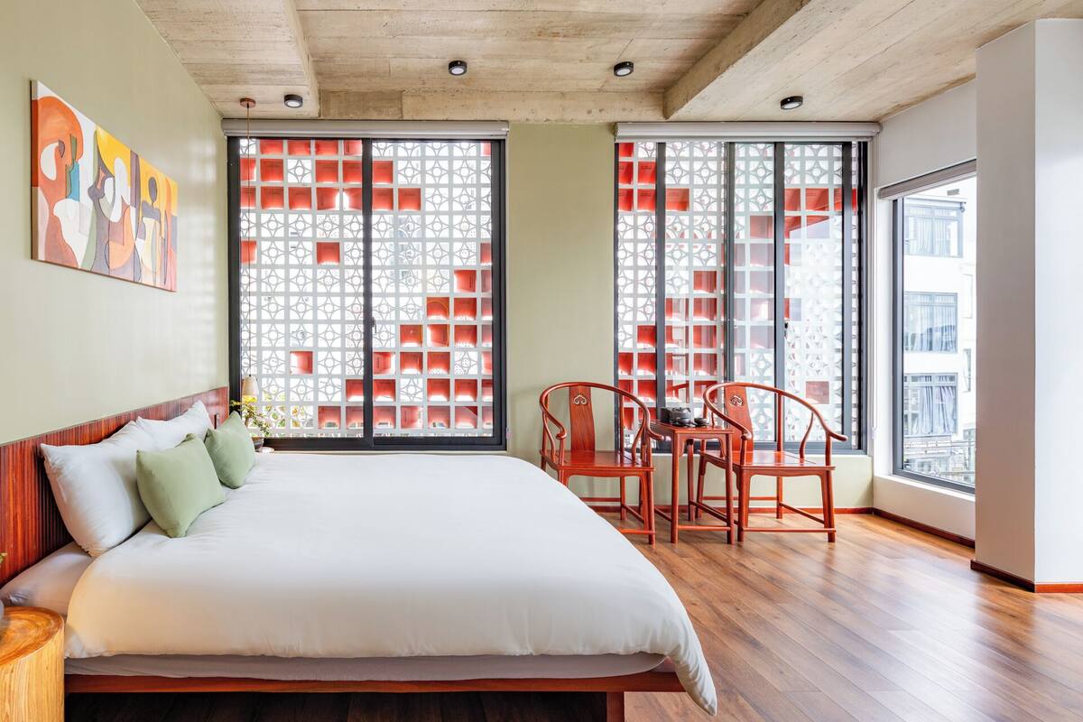 Une chambre moderne avec un grand lit, un plancher en bois et une vue sur l’extérieur, visible à travers une fenêtre agrémentée d’un paravent décoratif.