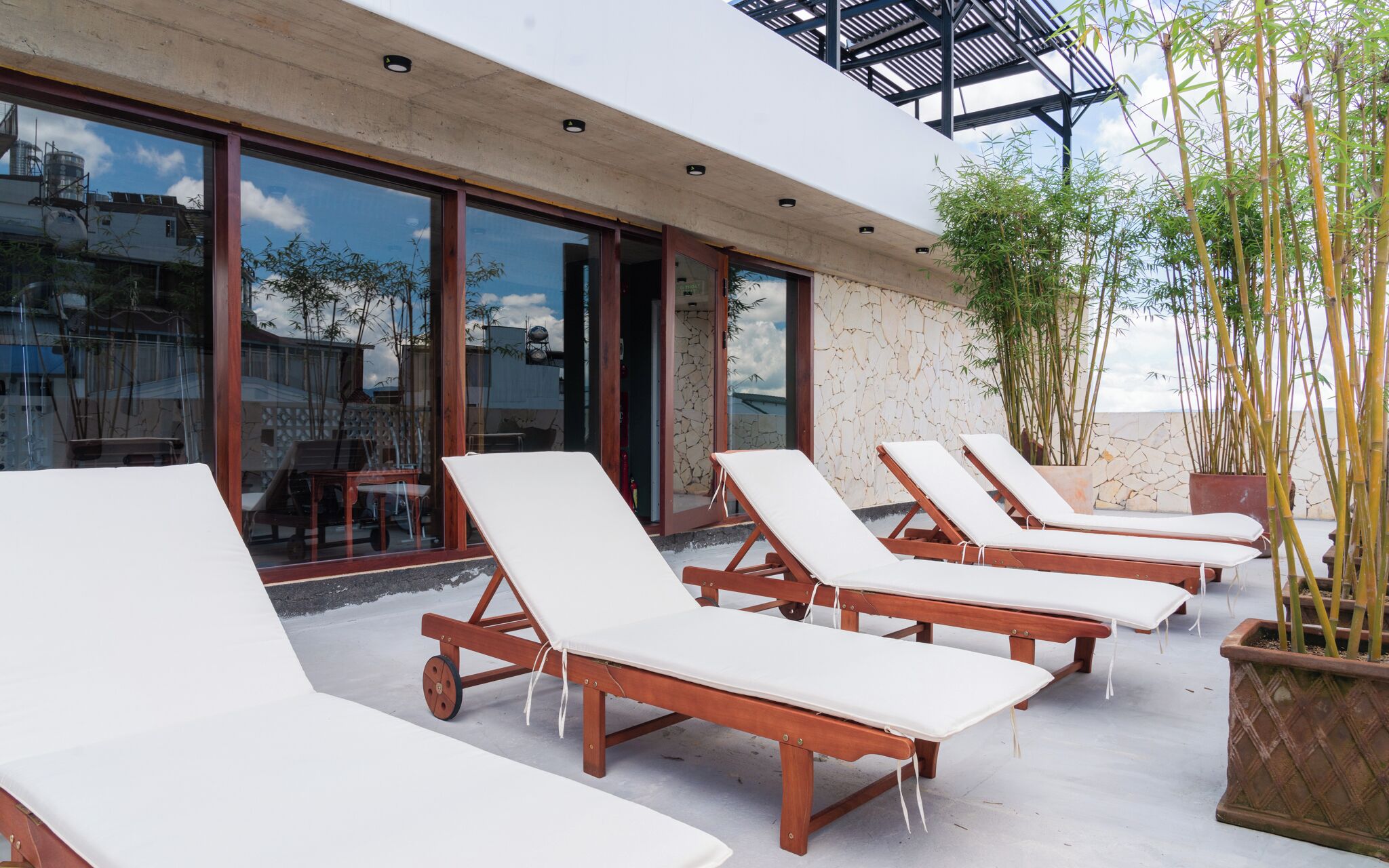 A row of wooden lounge chairs with white cushions on a rooftop terrace, surrounded by large windows and potted bamboo plants.