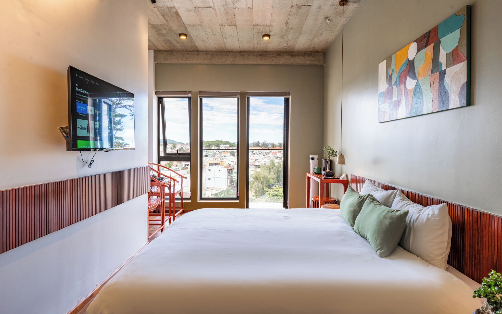 A hotel room with a large bed, a TV mounted on the wall, a painting, and a view of the city through the windows.