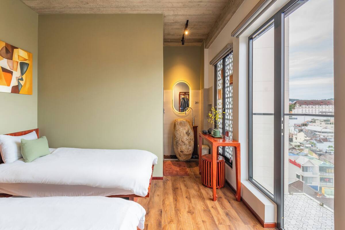 Une chambre d’hôtel avec deux lits, un plancher en bois, une grande fenêtre donnant sur la ville et un tableau au mur.