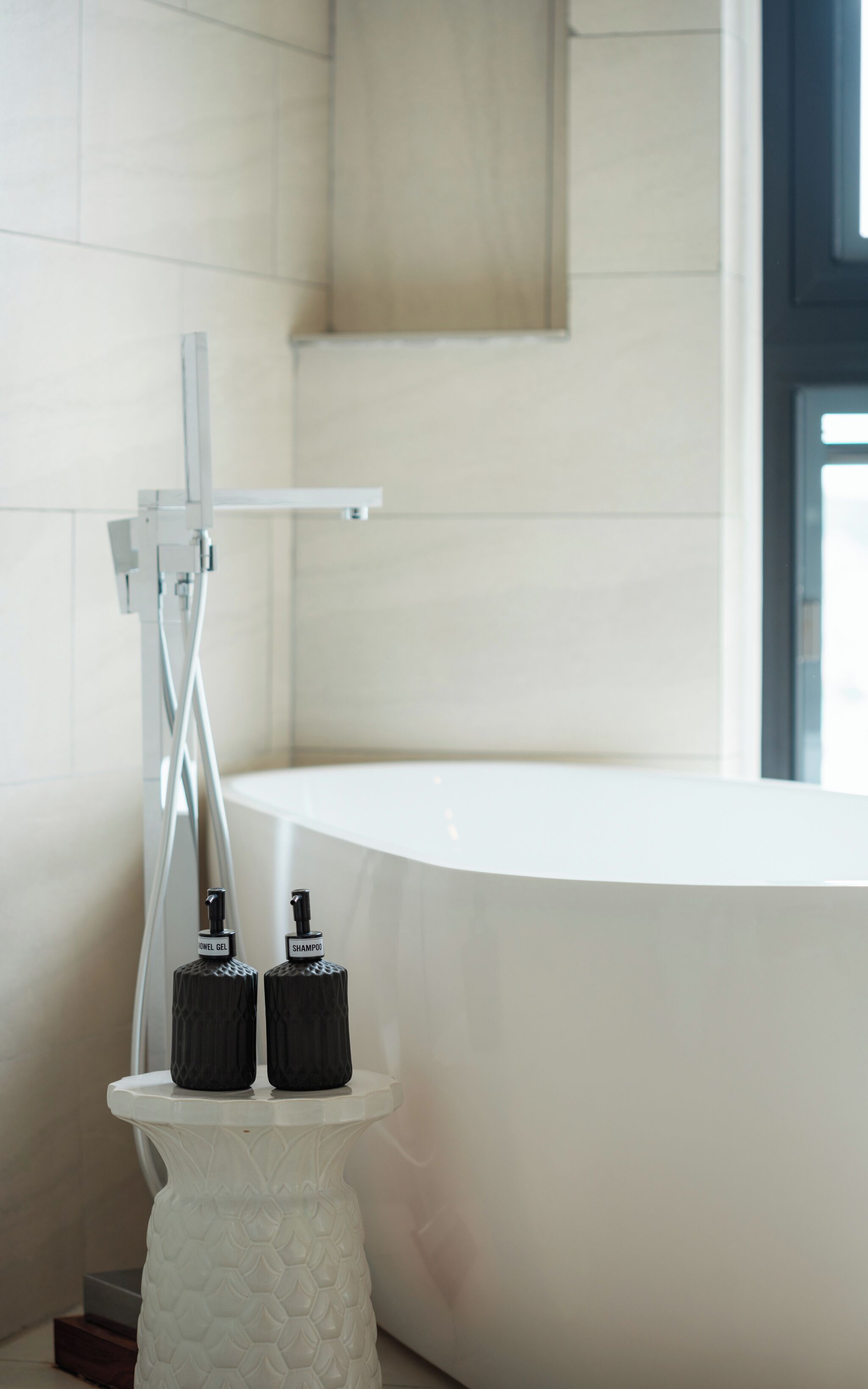 Un baño moderno con una bañera blanca, una ducha y dos botellas negras sobre un pedestal blanco.