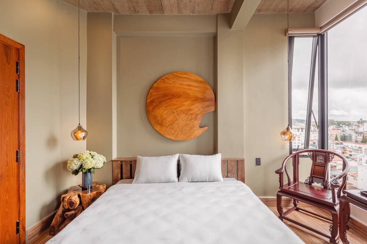 Une chambre avec un grand lit, une œuvre d’art murale en bois, une chaise en bois et une vue sur la ville par une grande fenêtre.