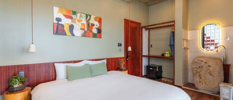 A bedroom with a large bed, a wooden headboard, a wall-mounted abstract painting, a wooden nightstand with a plant, a mirror, and a stone sink.
