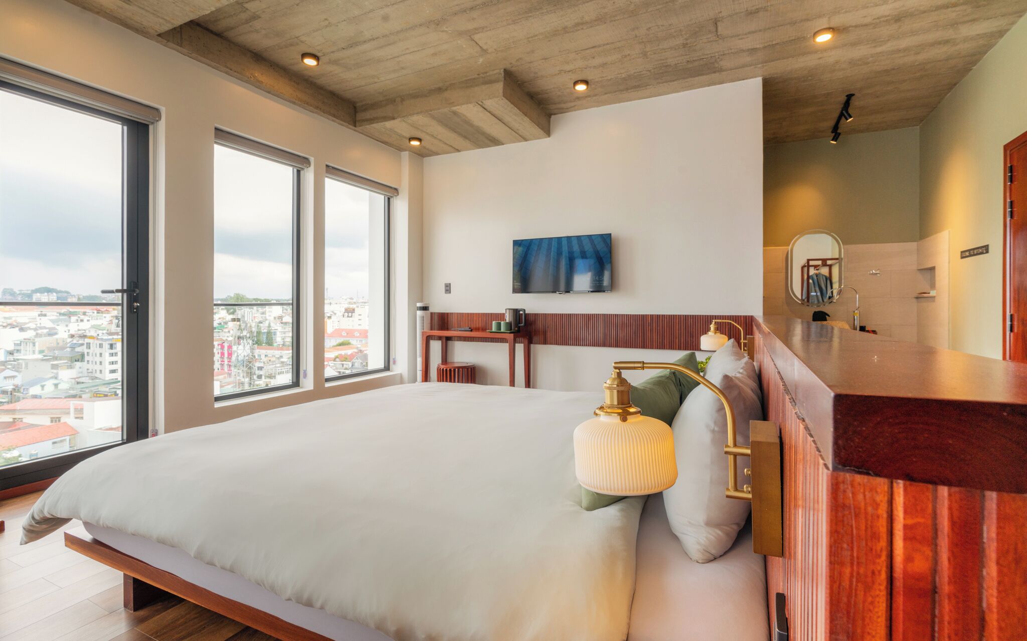 A modern hotel room with a large bed, a wooden headboard, a TV mounted on the wall, a small desk, and a bathroom with a mirror and sink.