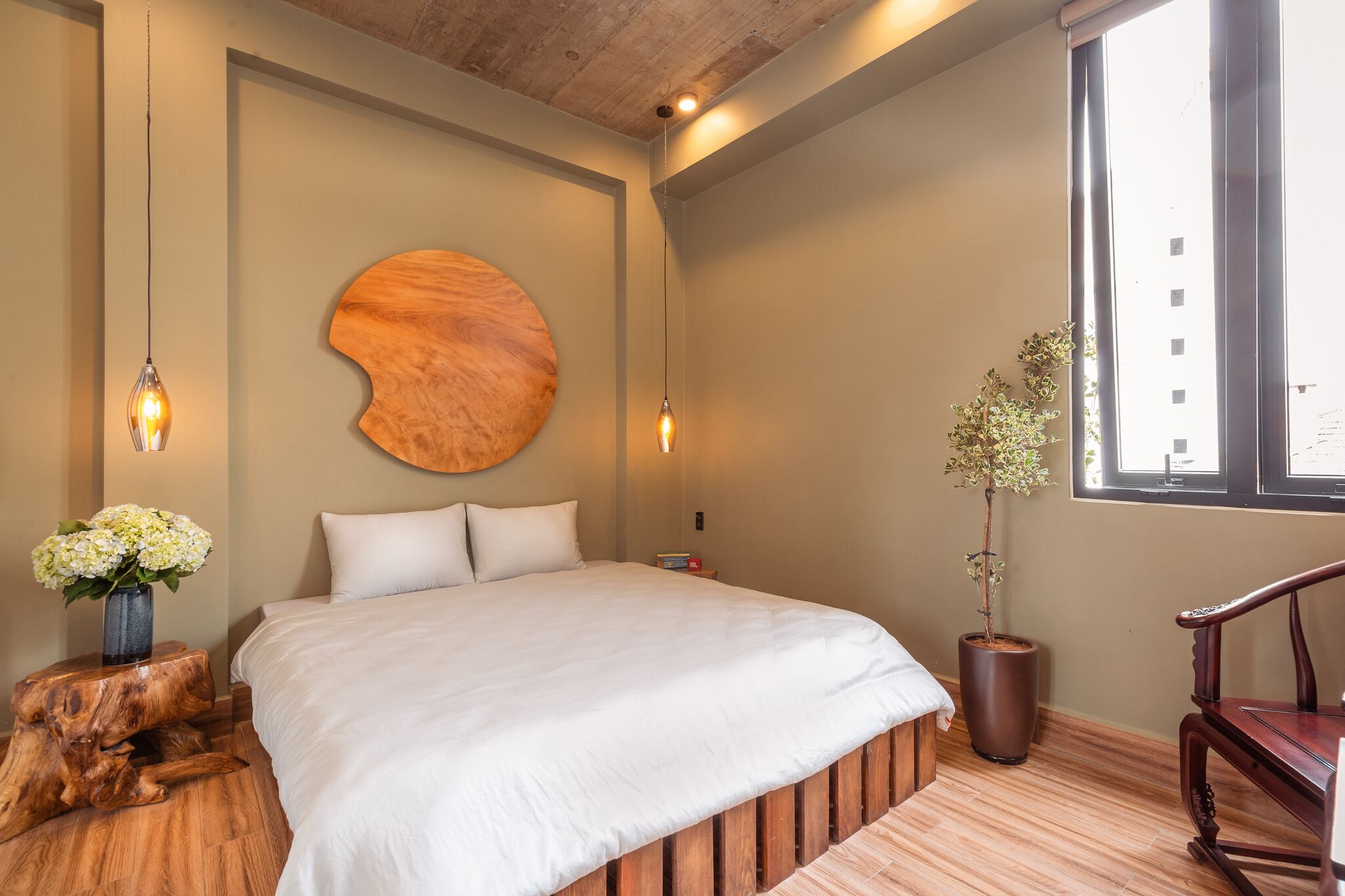 Une chambre moderne avec un grand lit, une tête de lit en bois, une décoration murale ronde en bois, une plante en pot et une chaise en bois.
