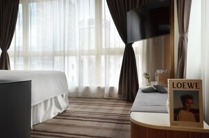 Habitación de hotel con cama, televisor, mesita de noche con un libro titulado 'LOEWE' y ventana con cortinas translúcidas.