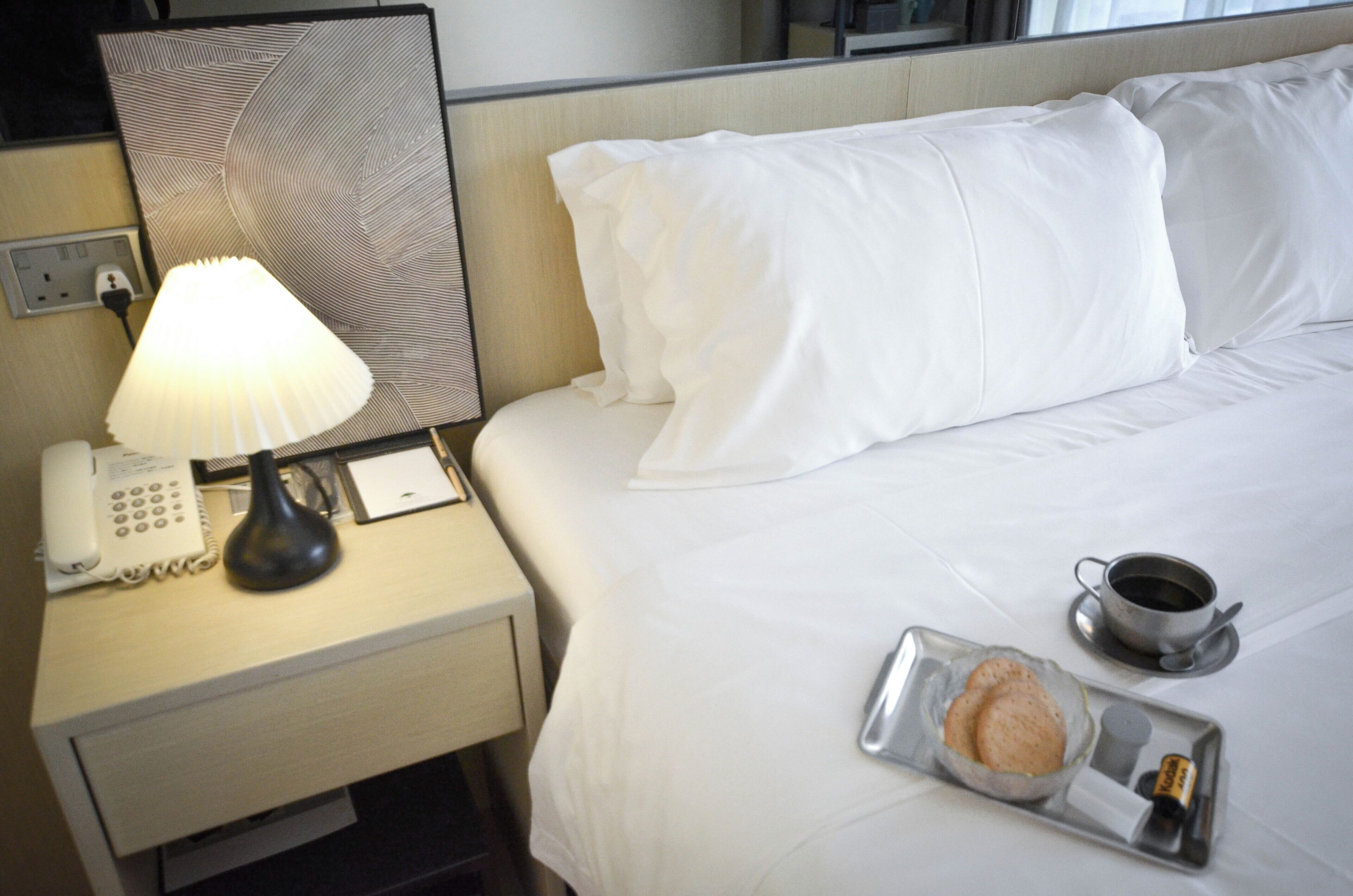 Une chambre d’hôtel avec un lit bien fait, une lampe de chevet, un téléphone, une tasse de café et un plateau avec un muffin et une bouteille.