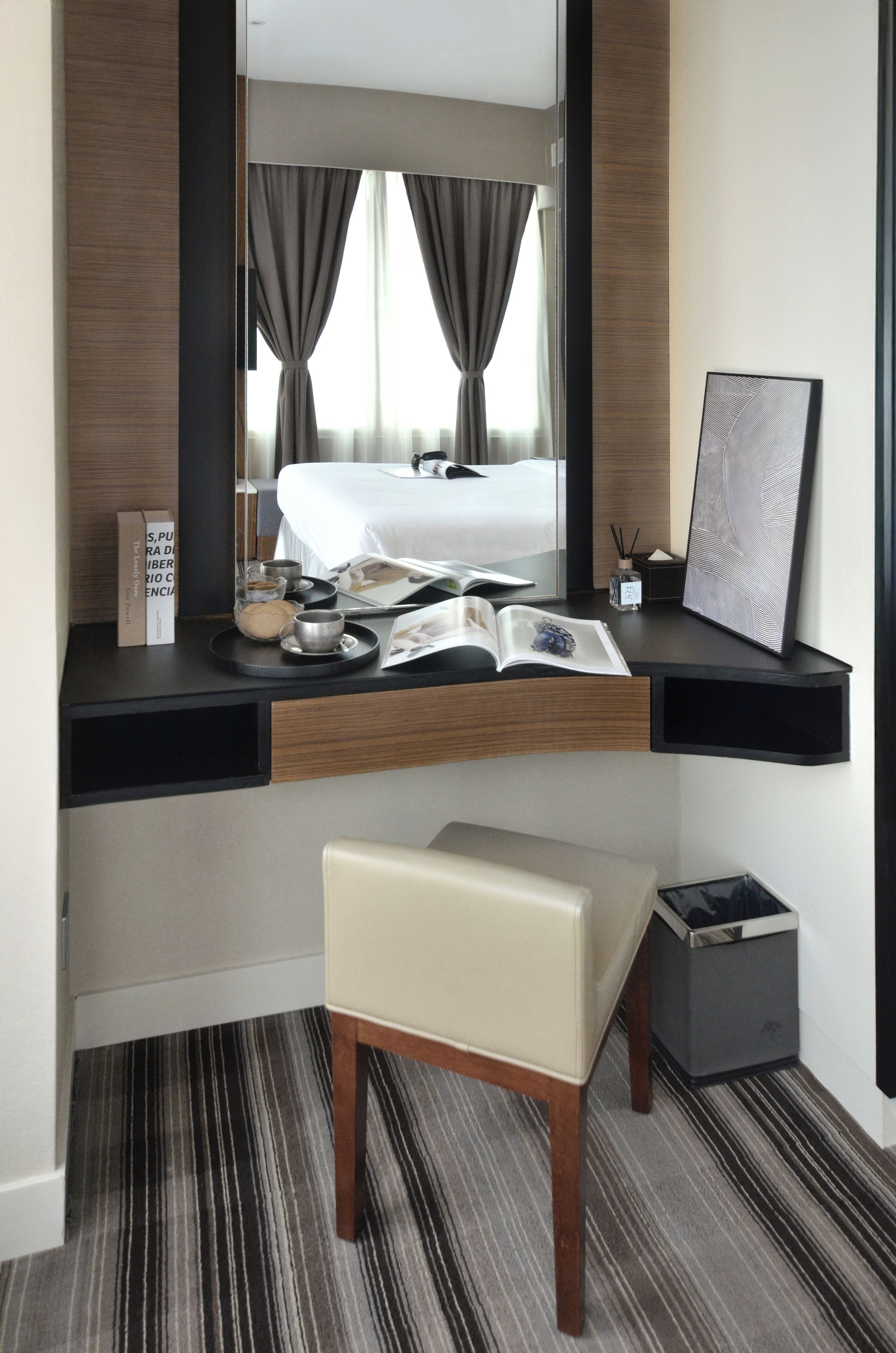 A modern hotel room with a built-in desk, a comfortable chair, a small trash bin, and a bedside table with a book and a decorative item.