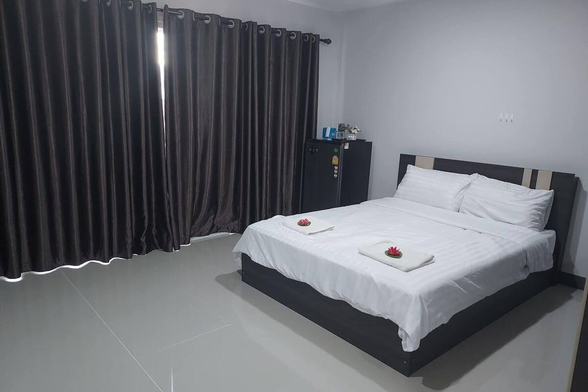 Una cama bien tendida con sábanas blancas y dos flores rojas, un armario oscuro y una ventana grande con cortinas oscuras.