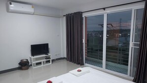 A hotel room with a flat-screen TV on a white stand, a large sliding glass door with curtains, and a bed with neatly folded white towels. - Thana Apartment (Si Racha)