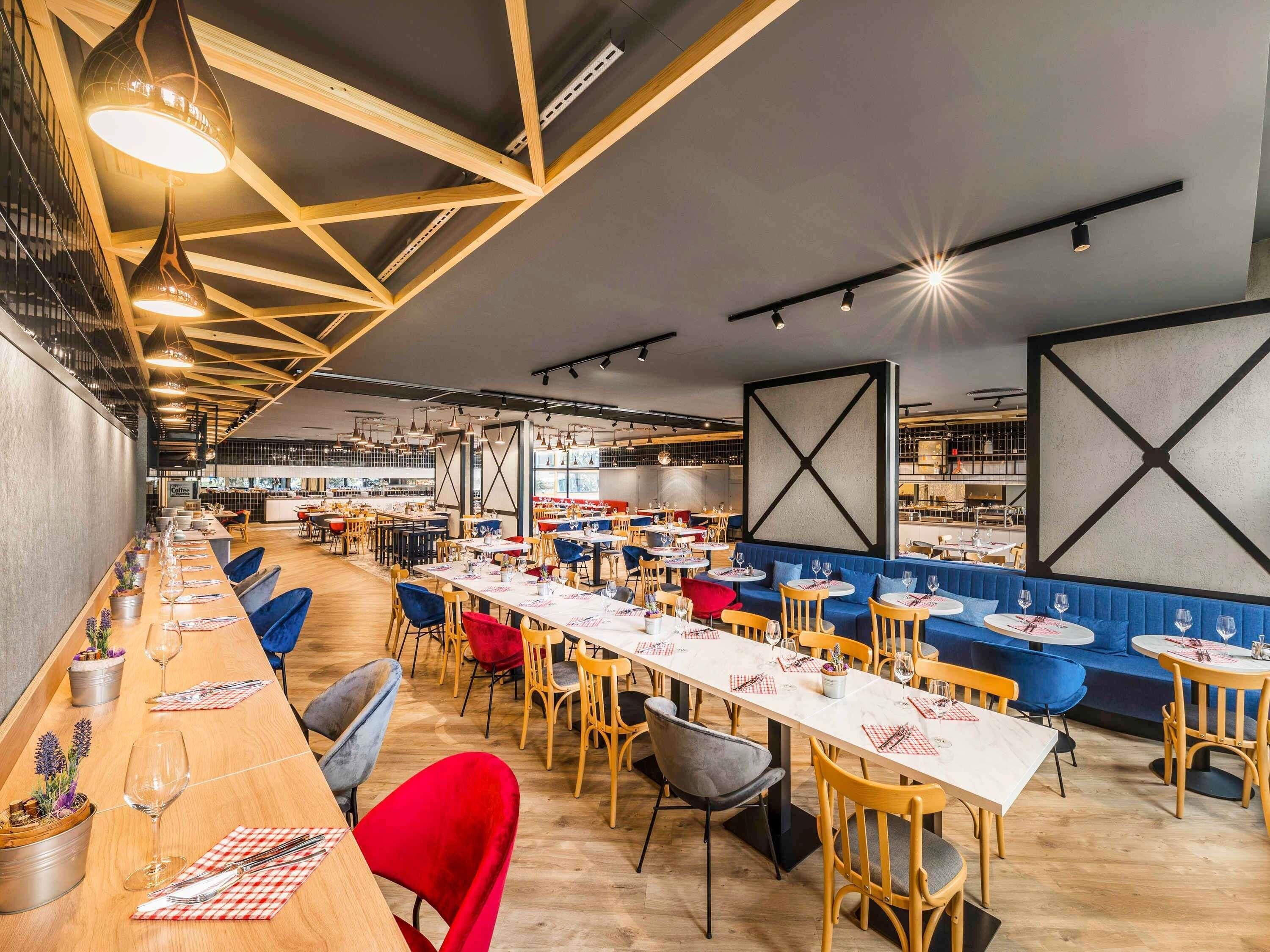 A modern restaurant with long communal tables, individual booths, and a high ceiling with exposed beams.