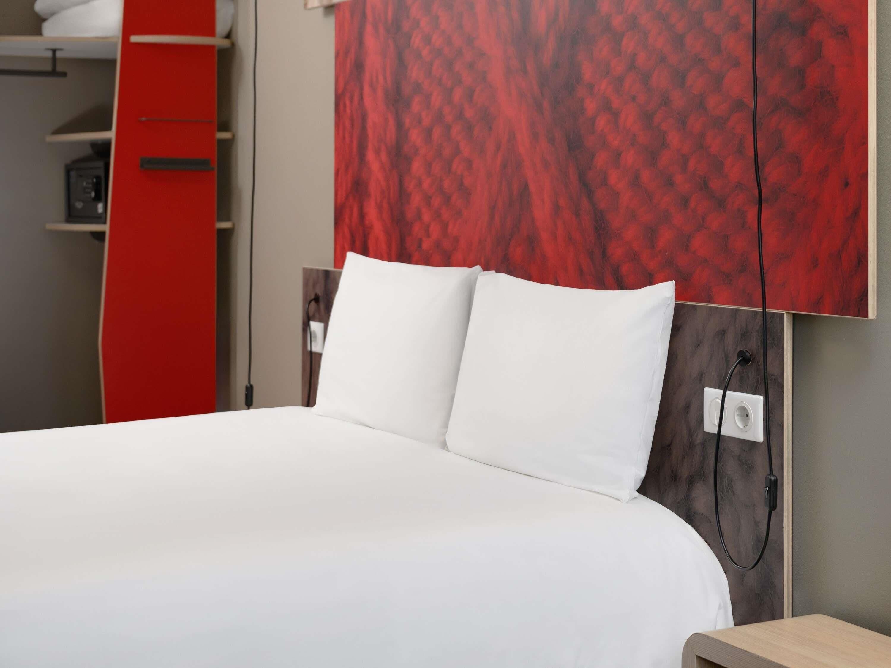 A neatly made bed with white bedding, a red wardrobe, and a wall with a red abstract pattern.