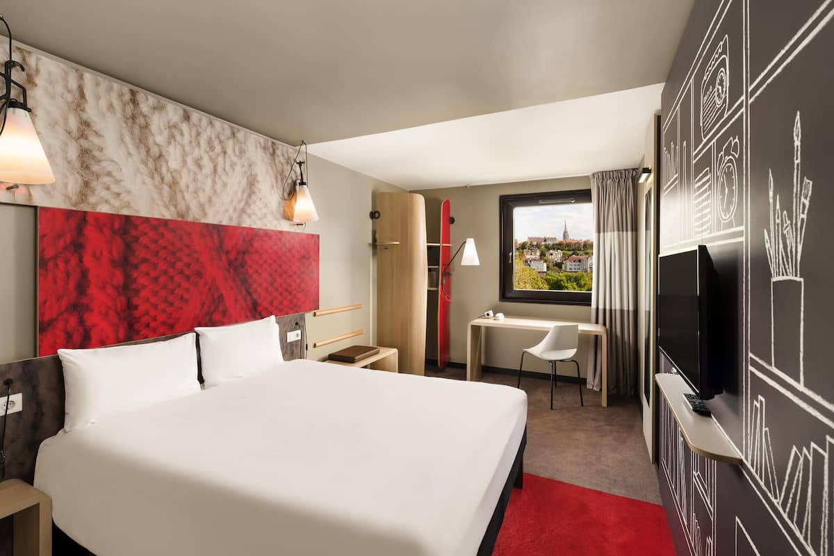 Habitación de hotel con una cama grande, un escritorio, una silla, un televisor y una ventana con vista a la ciudad.