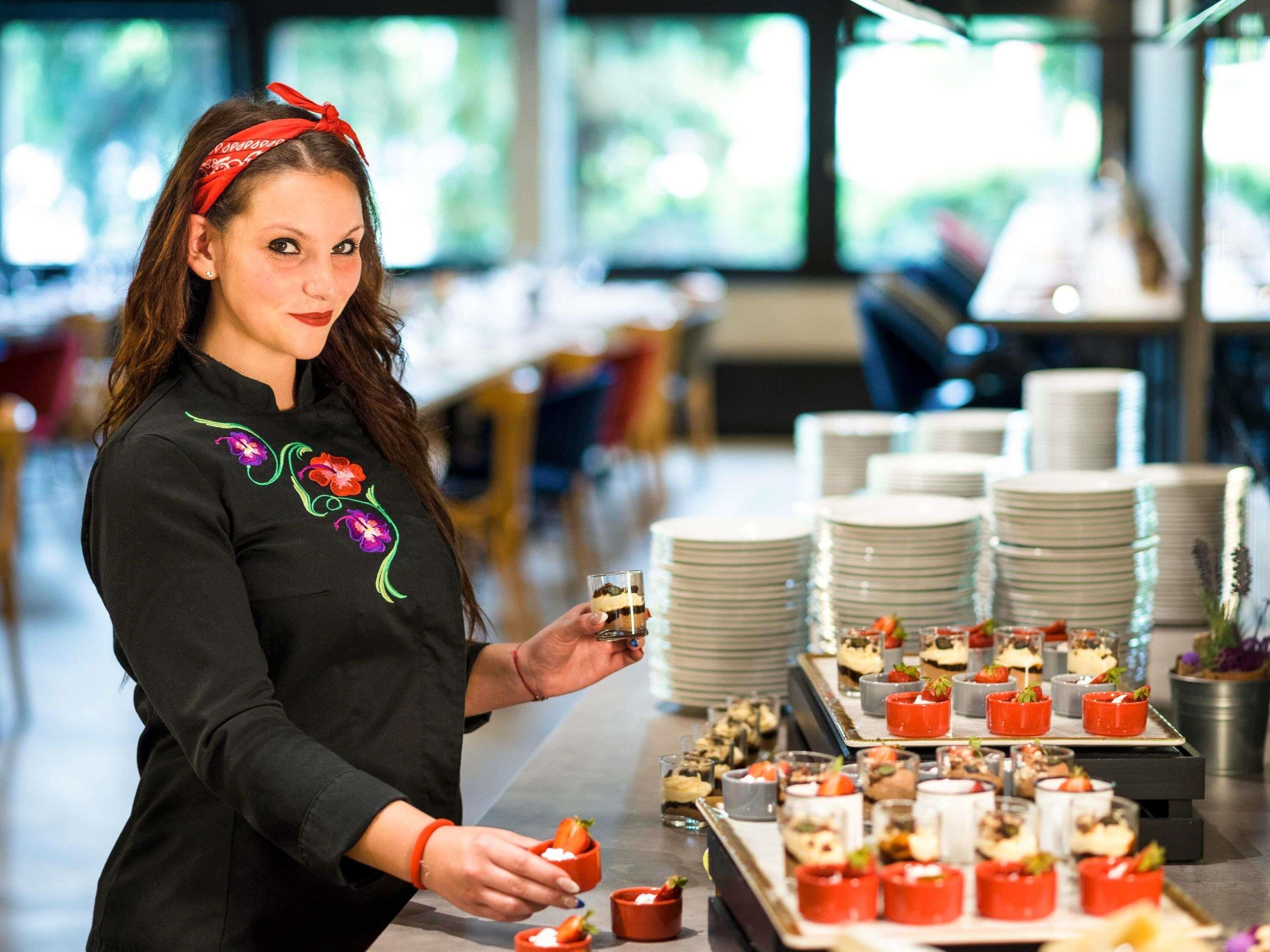 Una persona sistema alimenti su un tavolo del buffet, sullo sfondo si vedono pile di piatti bianchi.