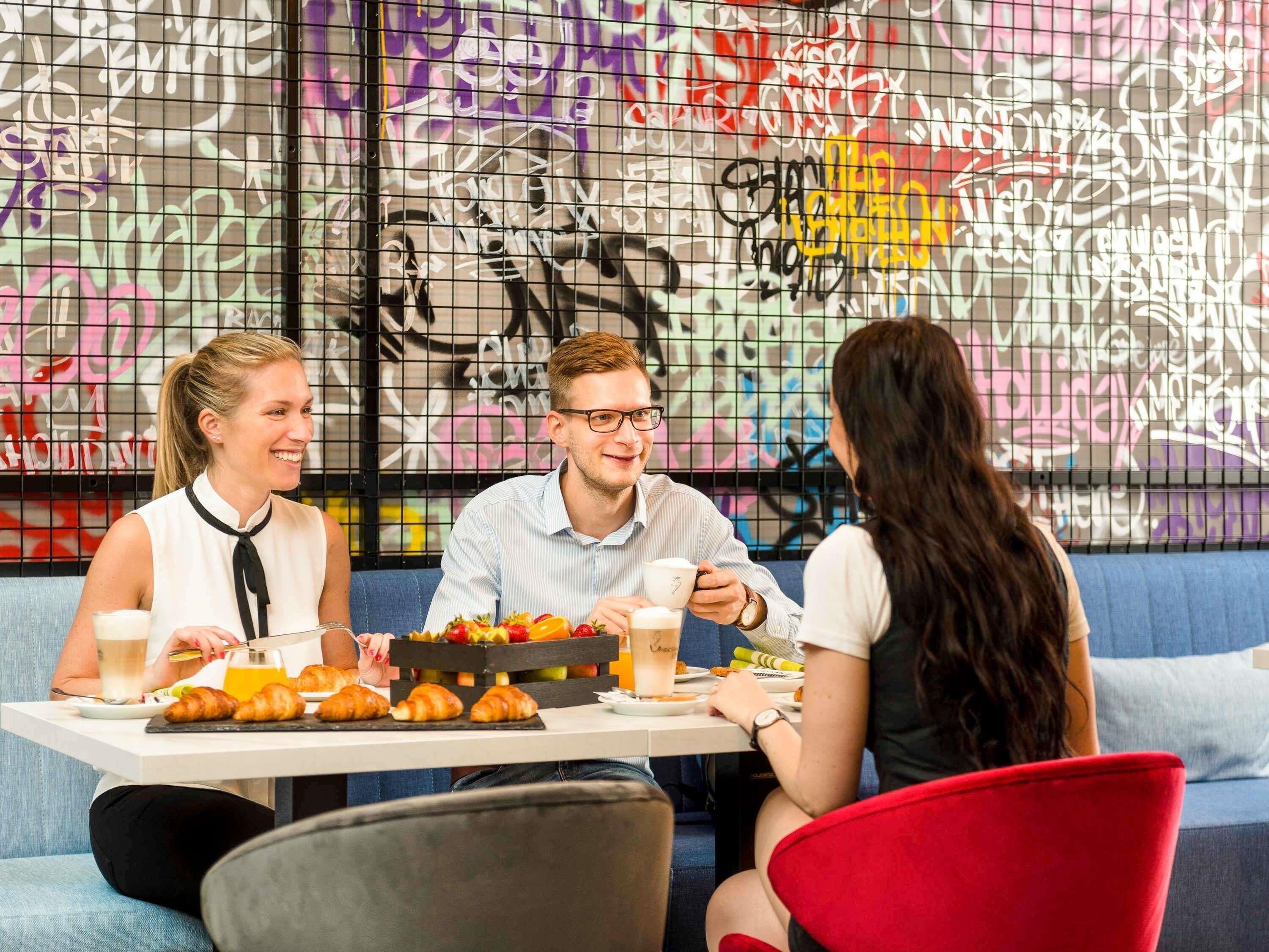 A group of people sitting at a table with food and drinks, in a room with graffiti on the wall.